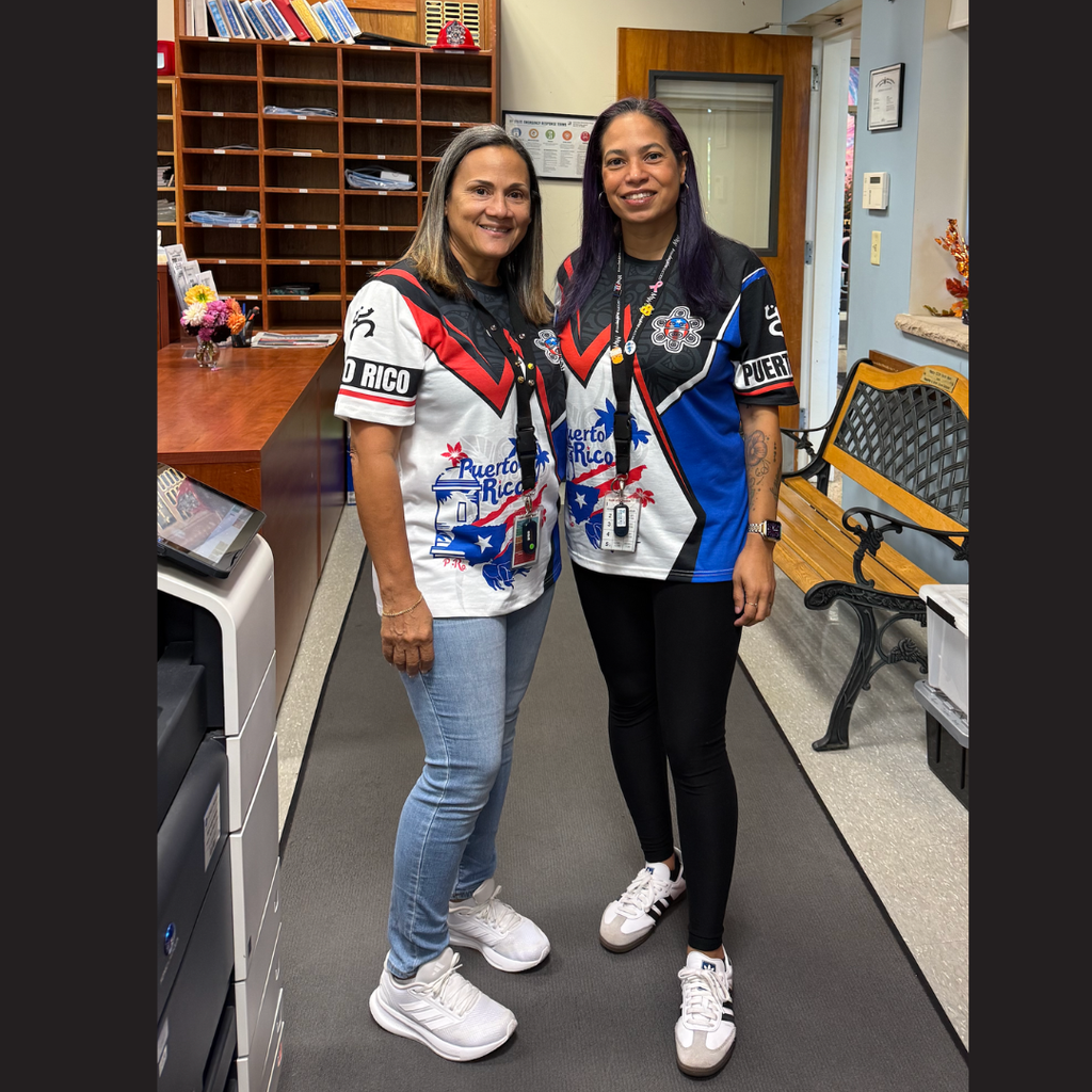 Two women in sports jerseys and sneakers stand in an office. One has purple hair; both wear name tags.