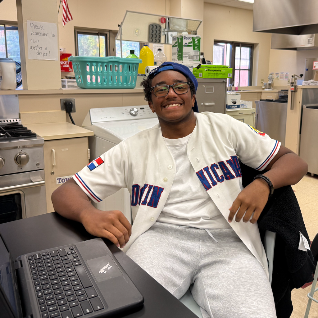 Man in baseball jersey sitting at a desk with a laptop, in a room with kitchen appliances.