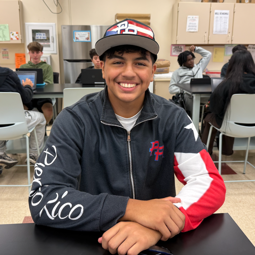 A student wearing a black and red jacket with "Puerto Rico" on the left arm, seated in a classroom.