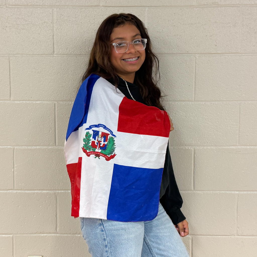 Person with long dark hair, glasses, and wearing a blue denim jacket holds a flag over shoulders.