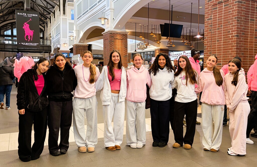 Several girls stand in a row wearing pink shirts and white pants, smiling and posing for a photo.