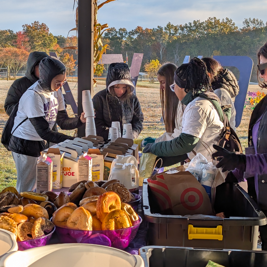 People serving food in bowls under a covered pavilion, surrounded by trees and a storage box.