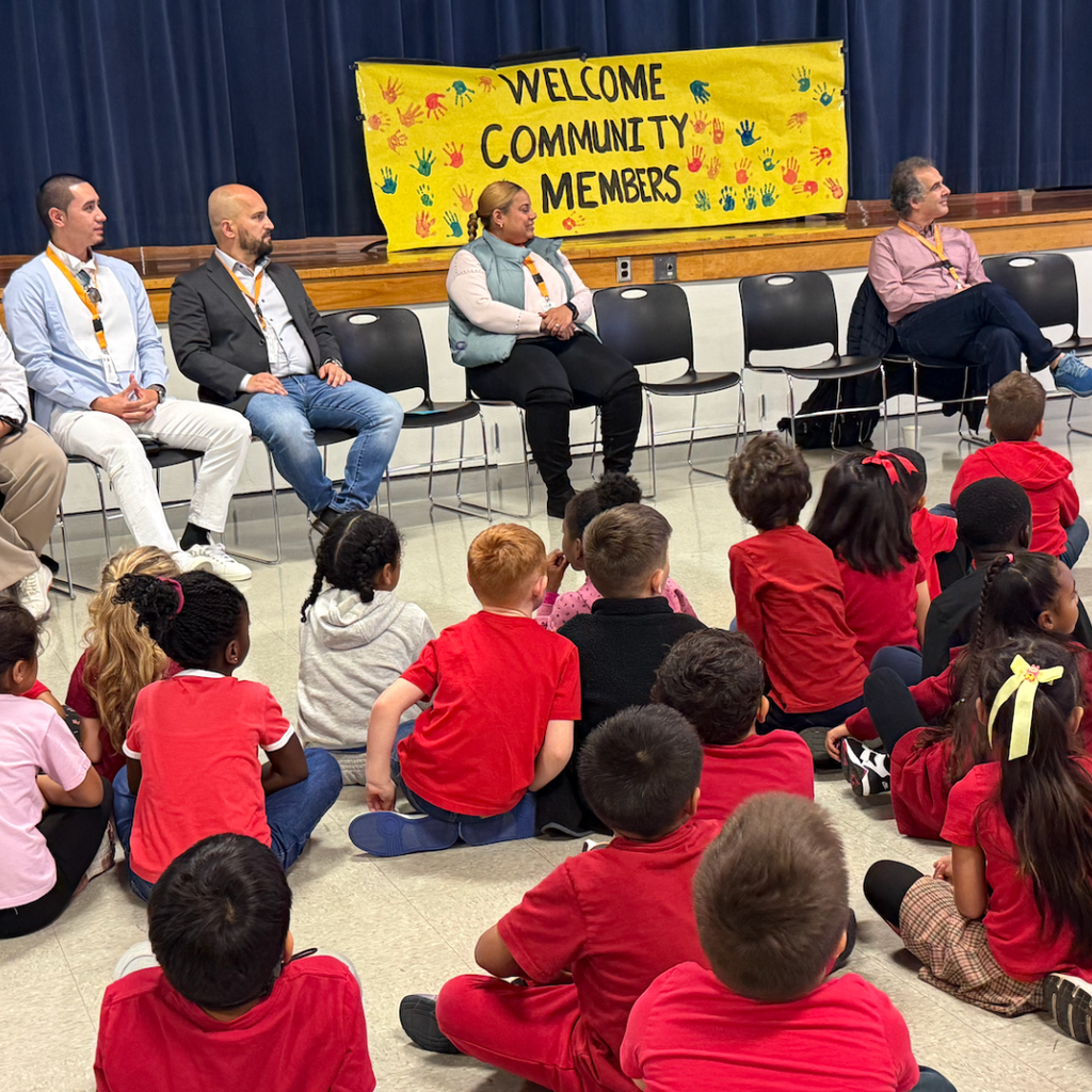 Group of adults and children seated in chairs. Banner above them reads "Welcome Community Members". Children wear red shirts.
