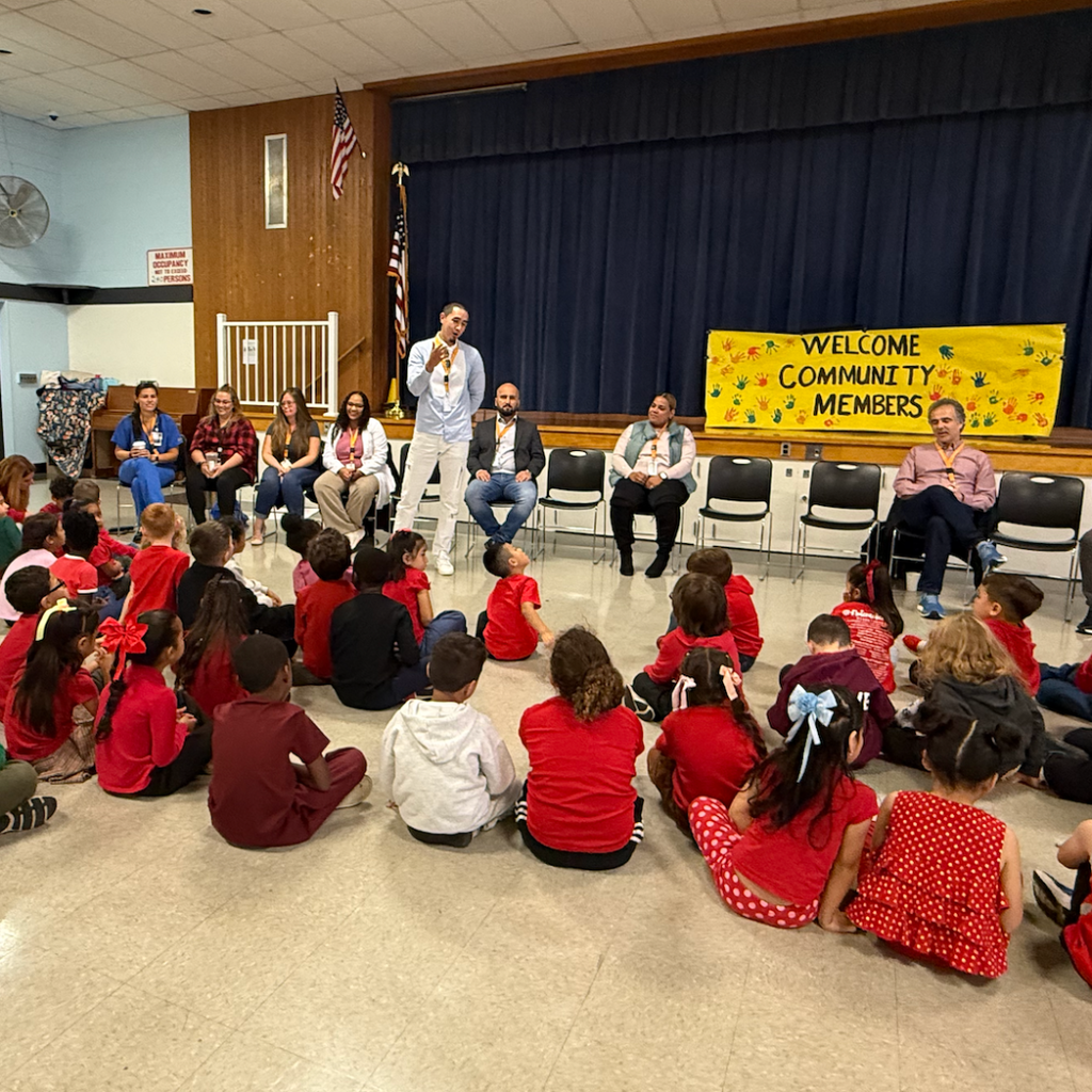 Inside, children in red and white sit in a circle. One adult stands with a microphone. A banner reads Welcome Community Members.