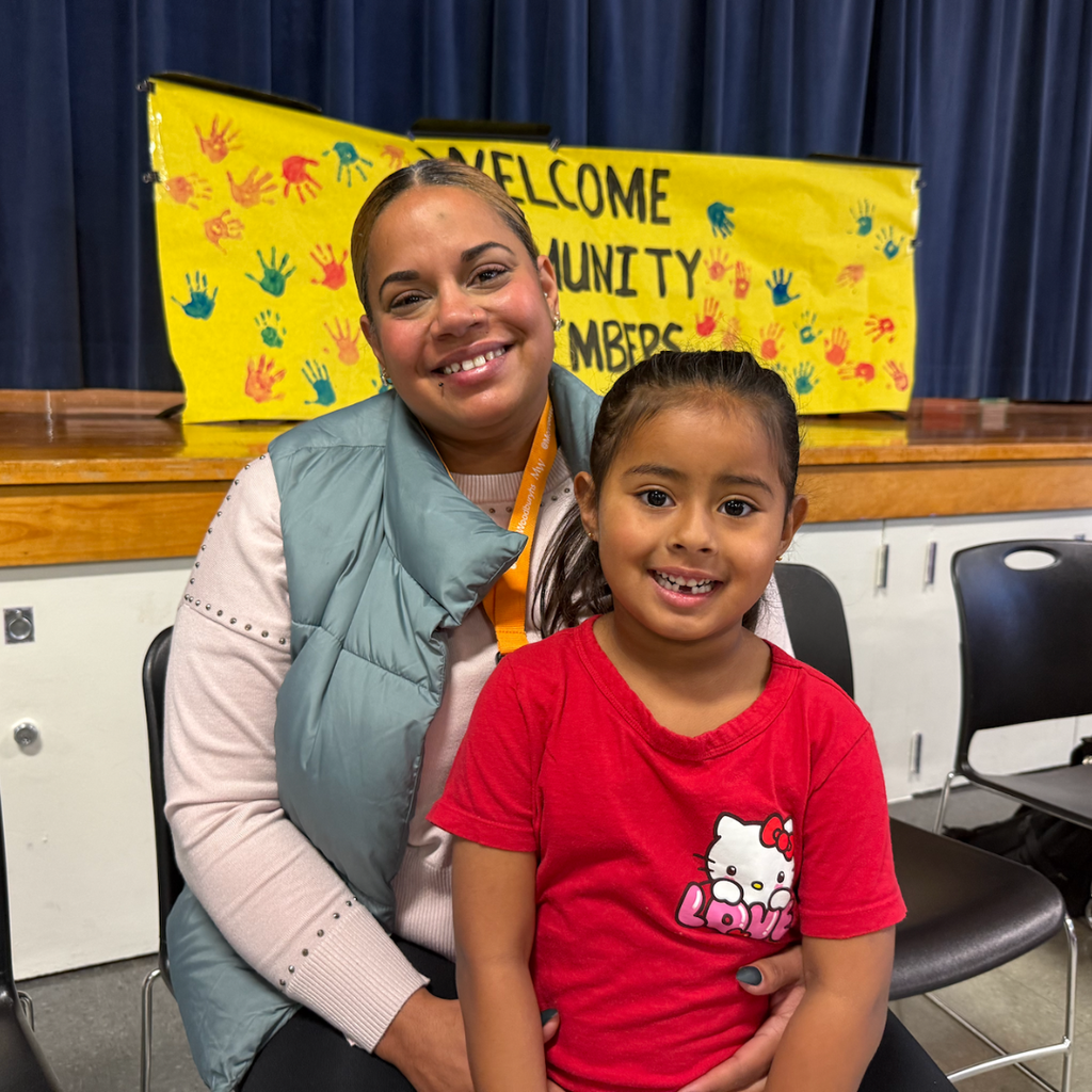 A woman and young girl sit side by side in chairs, smiling. A banner with handprints hangs in the background.