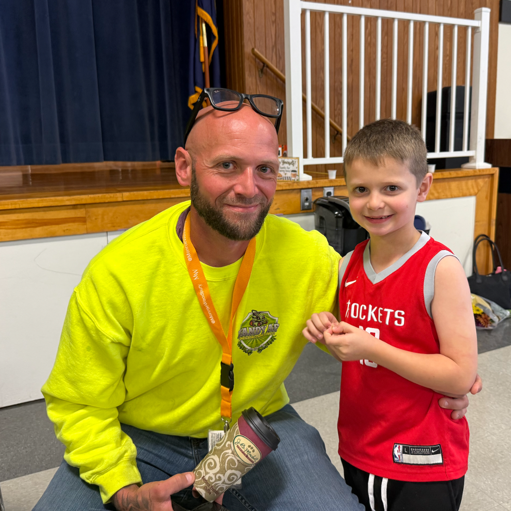 A man and a boy stand in a room. The man wears a yellow shirt and holds a cup. The boy wears a red jersey and smiles.