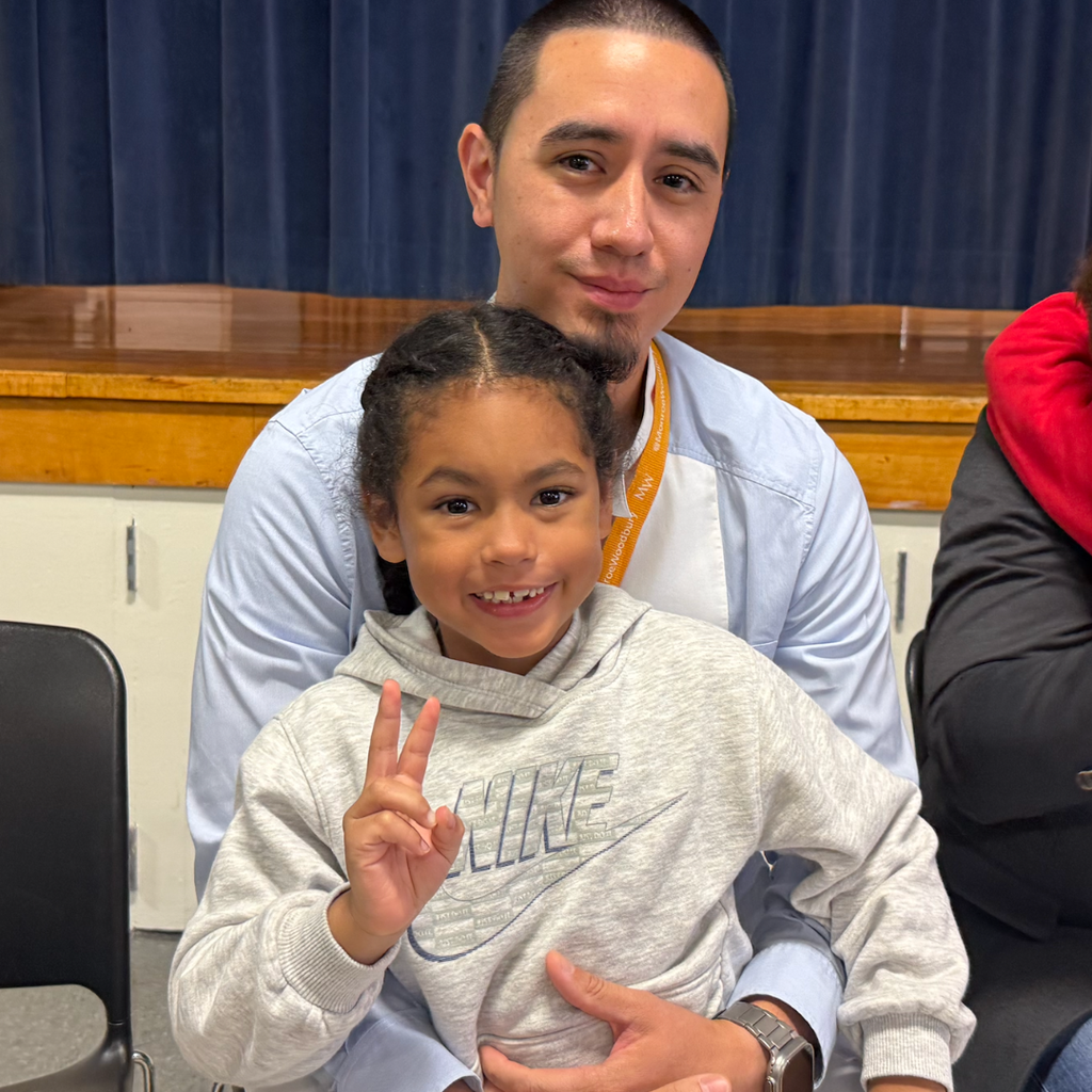 Man, girl, and another person in classroom. Girl in hoodie makes peace sign. Man smiles.
