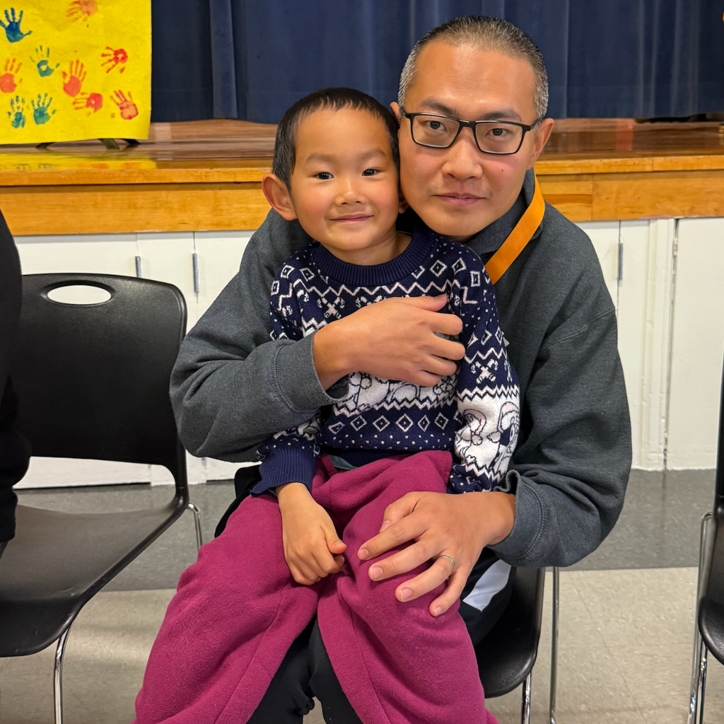 Man sitting in chair holding a child, wearing glasses. Child wearing blue sweater, purple pants, and smiling.