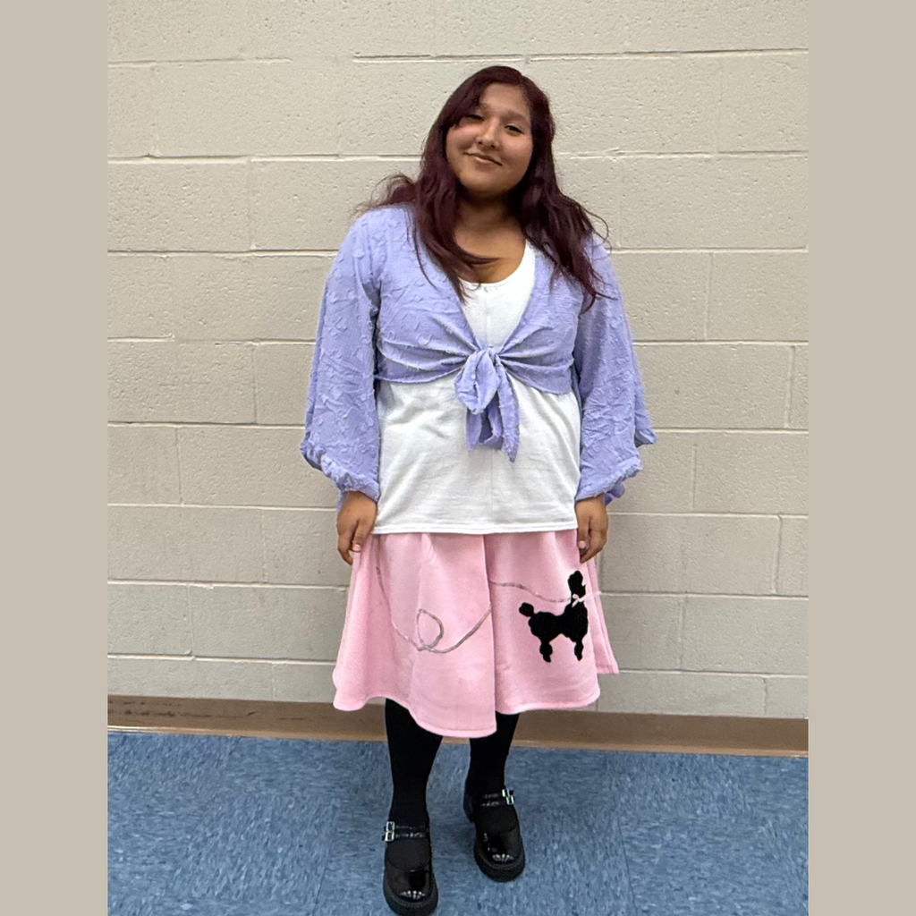 A person stands indoors, smiling at the camera, wearing a white top, pink skirt, and a purple cardigan.