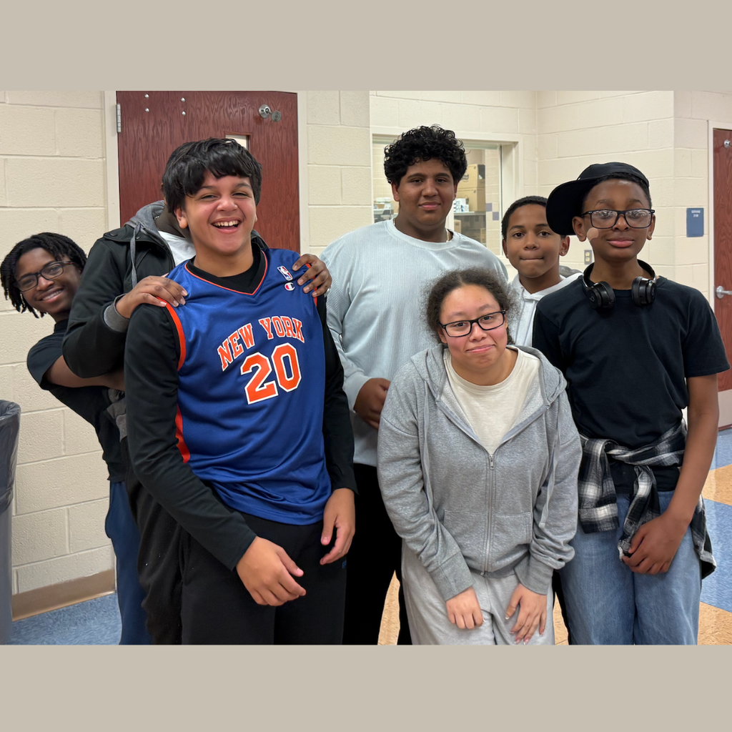 Six smiling students stand together in a hallway. A boy in a numbered jersey stands at the center.