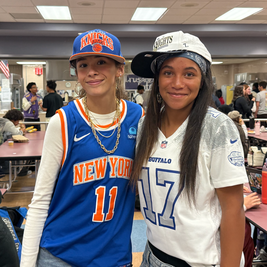 Two girls stand smiling with one wearing a blue and white jersey with the number 11.