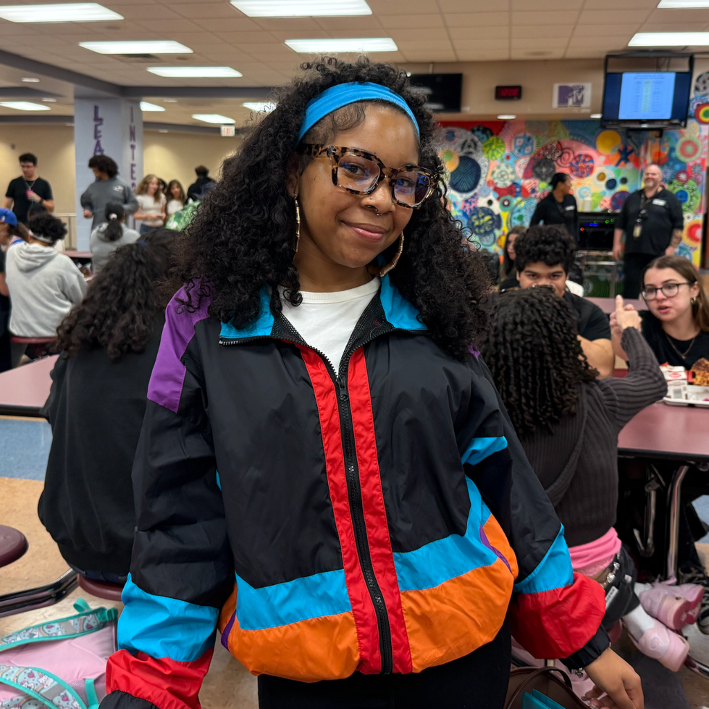 Woman in a multi-colored jacket stands in a school cafeteria; others sit at tables in the background.