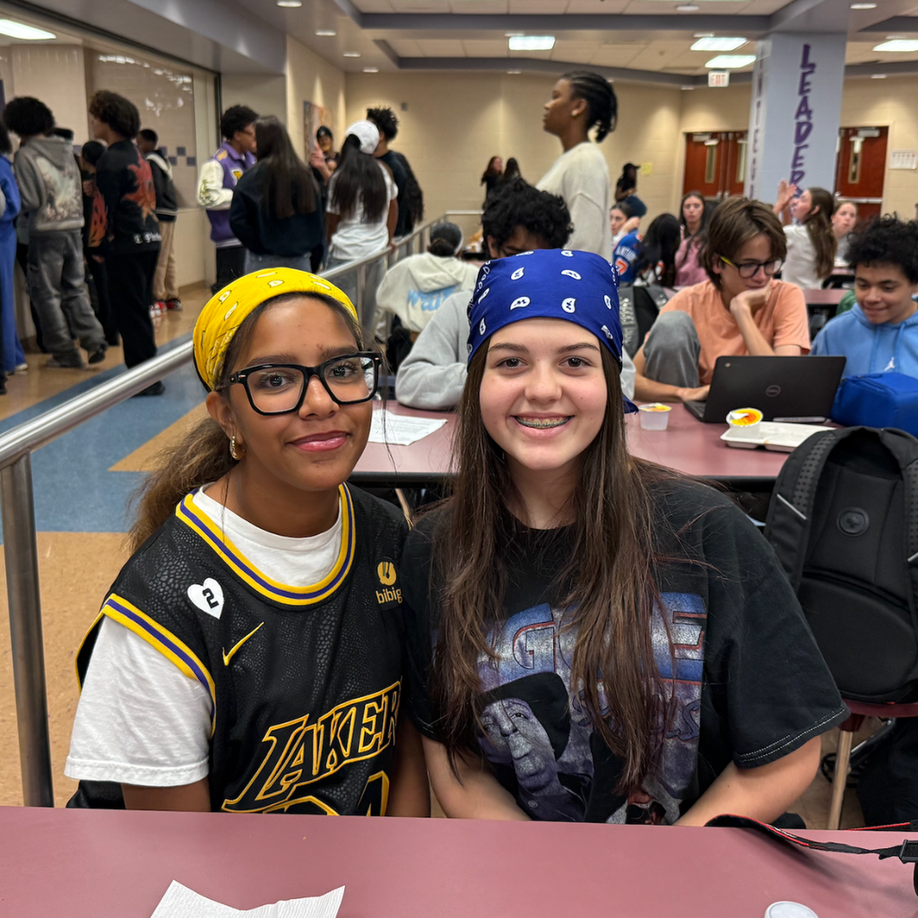 Two girls seated in a cafeteria; one wears a Lakers jersey, the other a blue bandana.