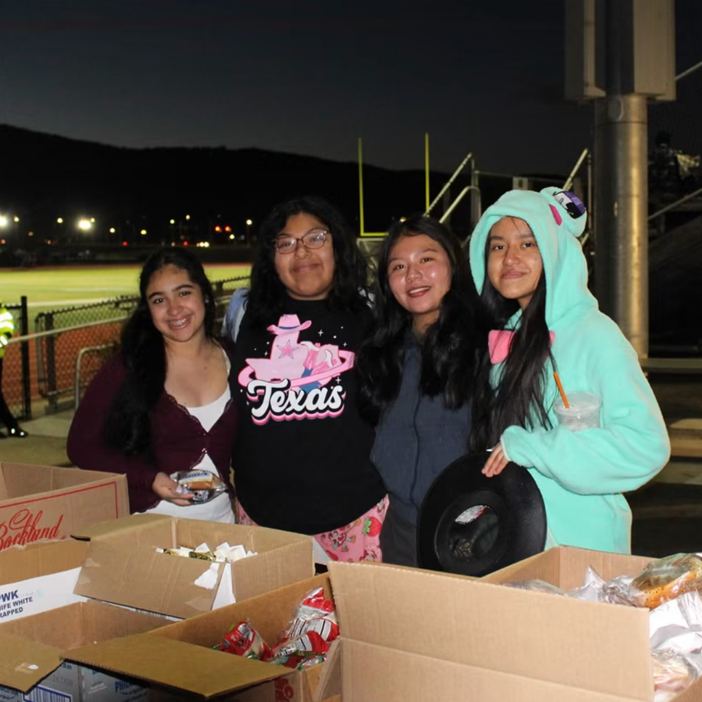 Four girls stand with boxes of food in a field with a stadium and mountains in the background.