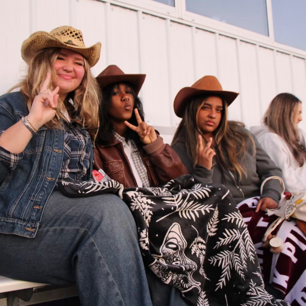 Four girls in cowboy hats seated on bleachers, two of them making peace signs, one covering mouth with hand.