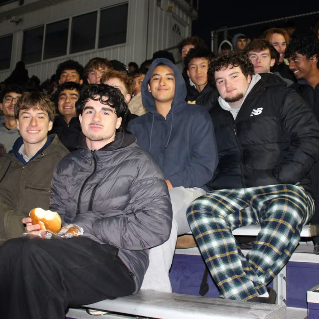 Group of male students sitting in bleachers at an outdoor event, one eating a hot dog.
