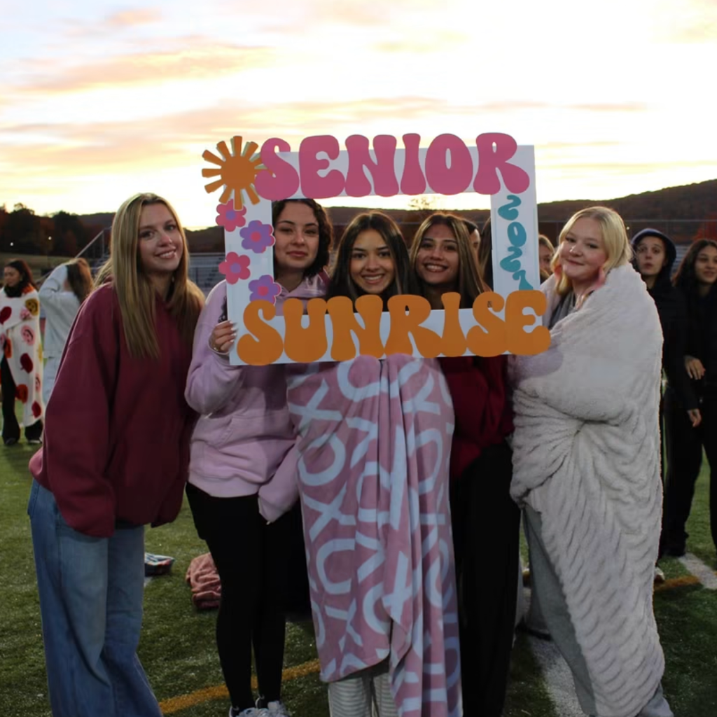 Students stand outside on a grassy field, wearing warm clothes. One holds a framed sign reading "Senior Sunrise".