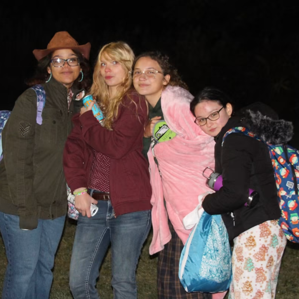 Four people stand together, two holding blankets, on a grassy field at night.