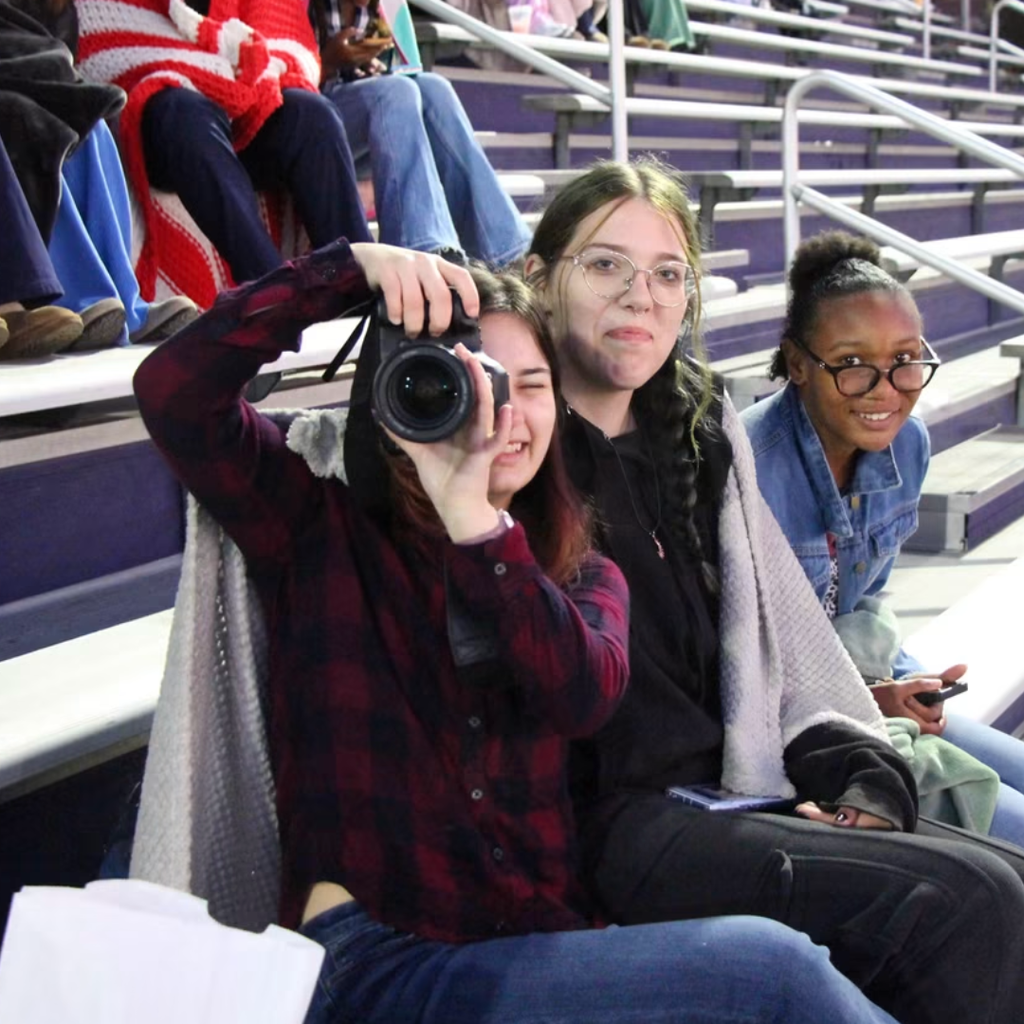 Three students seated on bleachers with one holding a camera and another holding a notebook.