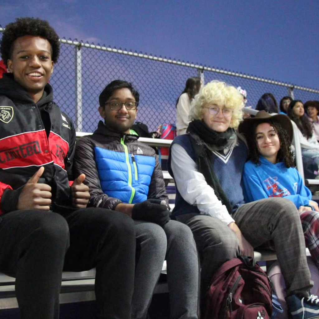 Four students sit together on a bleacher in the evening. They are wearing winter clothing and jackets.