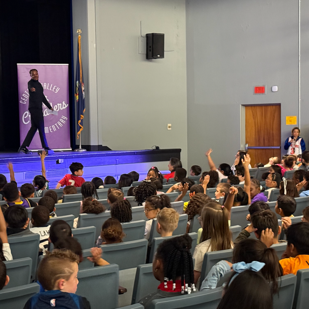 Auditorium with a stage, children seated in rows facing a banner, and a person walking on the stage.