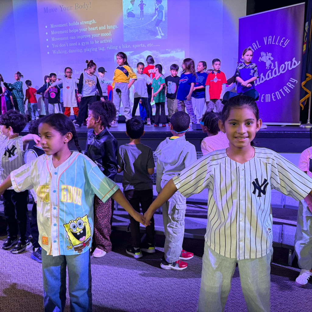 Children in baseball uniforms holding hands on stage with a projector screen in the background.