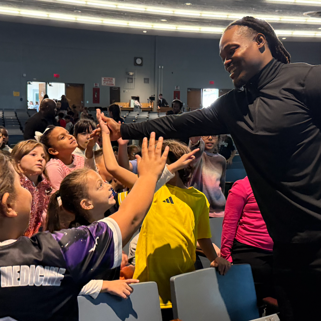 A man in a black top and kids in sports jerseys giving high-fives in a school auditorium.