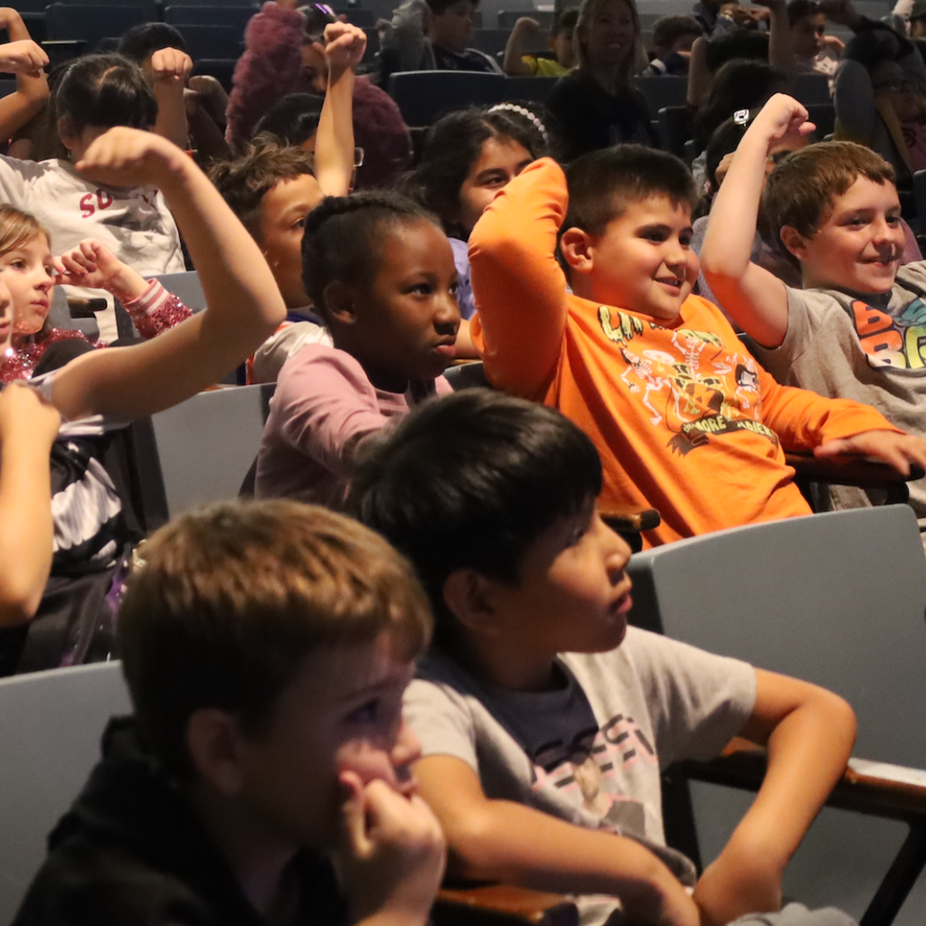 Children sit in chairs, looking at something in front of them, with some raising their hands.