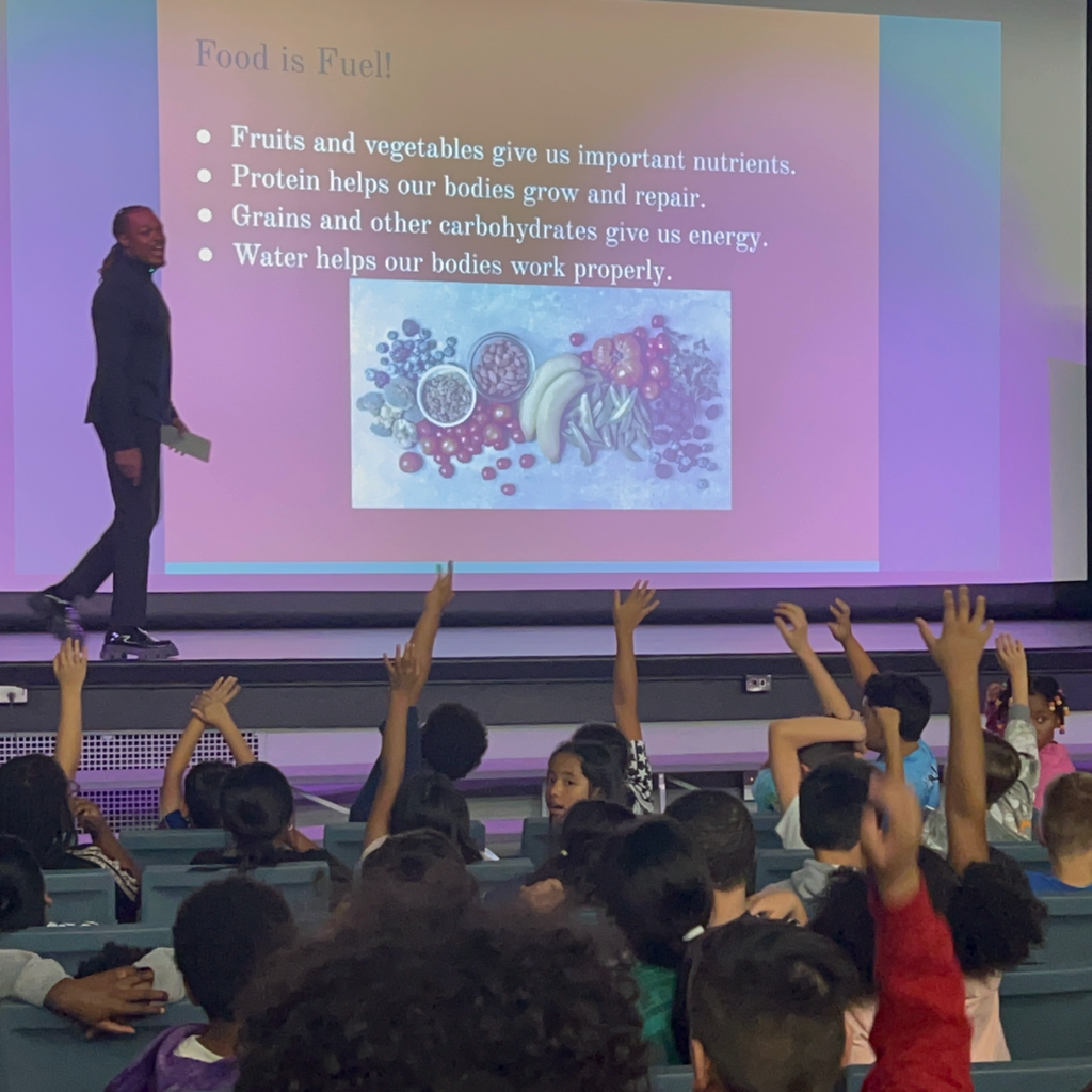 Teacher stands in front of screen with diagram of fruits and vegetables. Many children raise their hands.