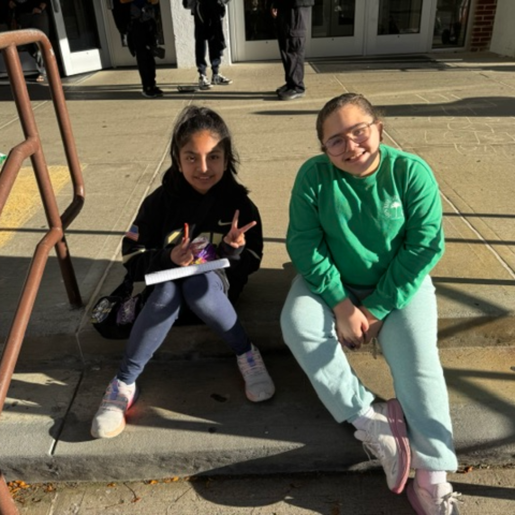Two young girls seated on steps, one with glasses and holding paper, the other in a green shirt.