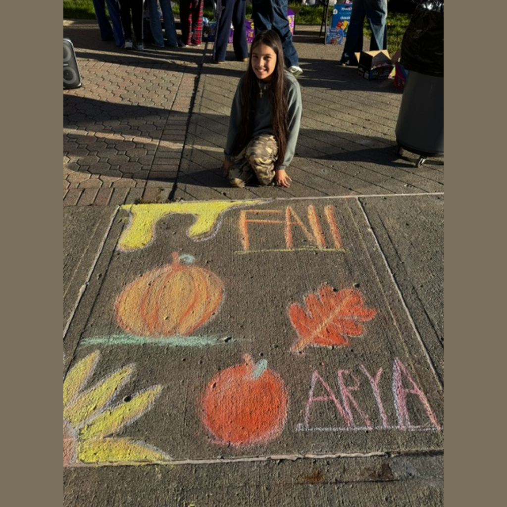A child sits on concrete. Pumpkins, leaves, and words are drawn with chalk on the ground.