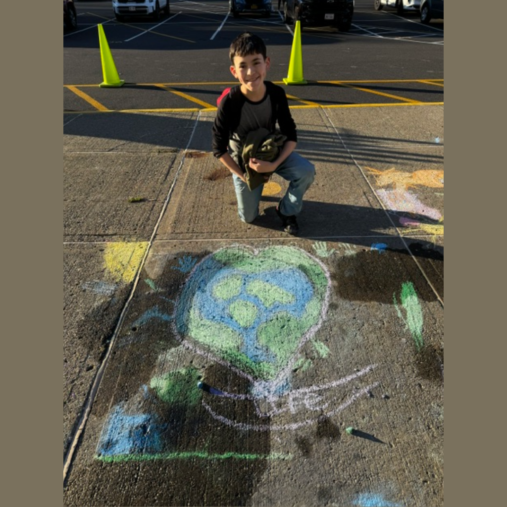 Child kneeling beside sidewalk chalk art. Background shows traffic cones and school parking lot.