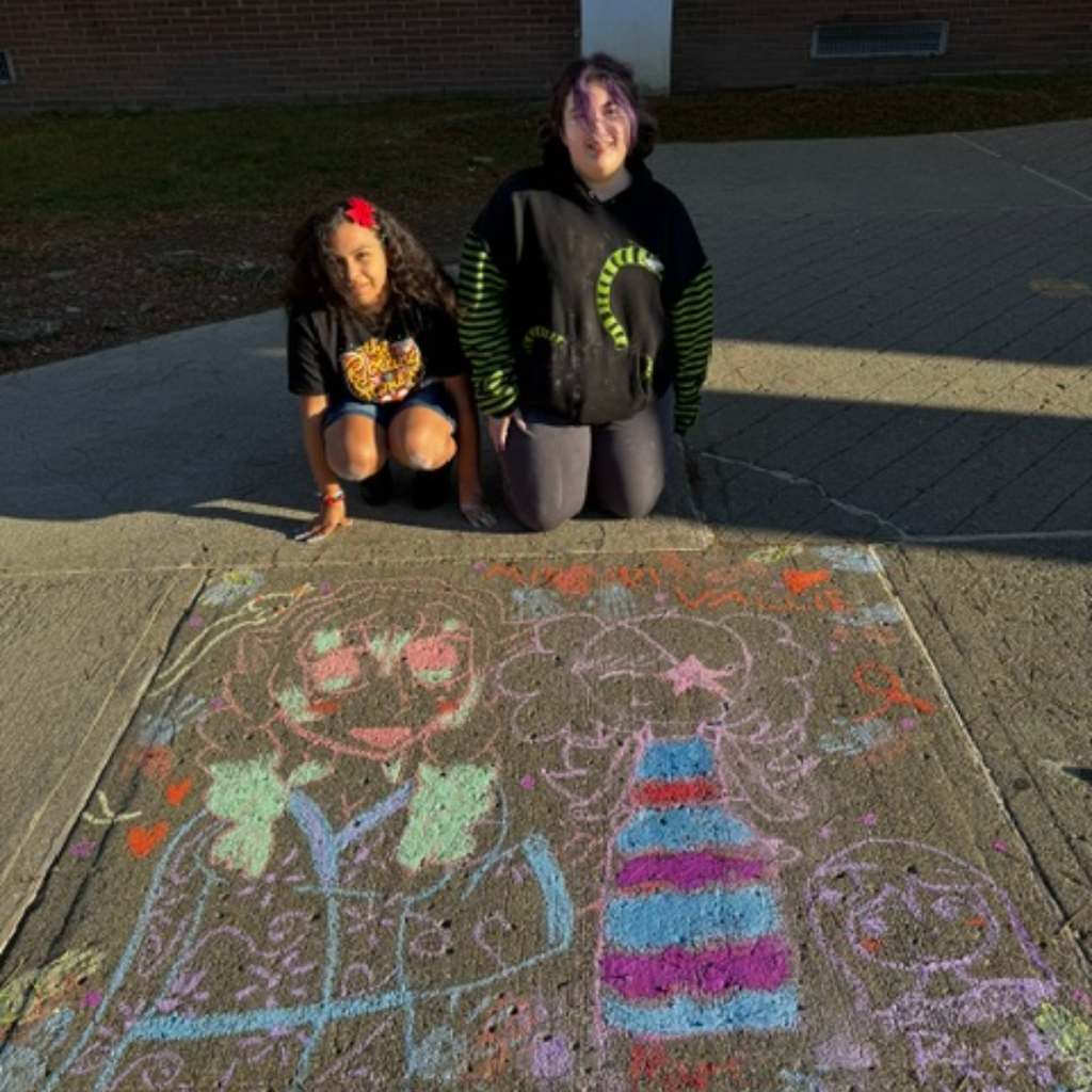 Two children crouch next to a chalk drawing of a bottle, faces, and buildings on a sidewalk.