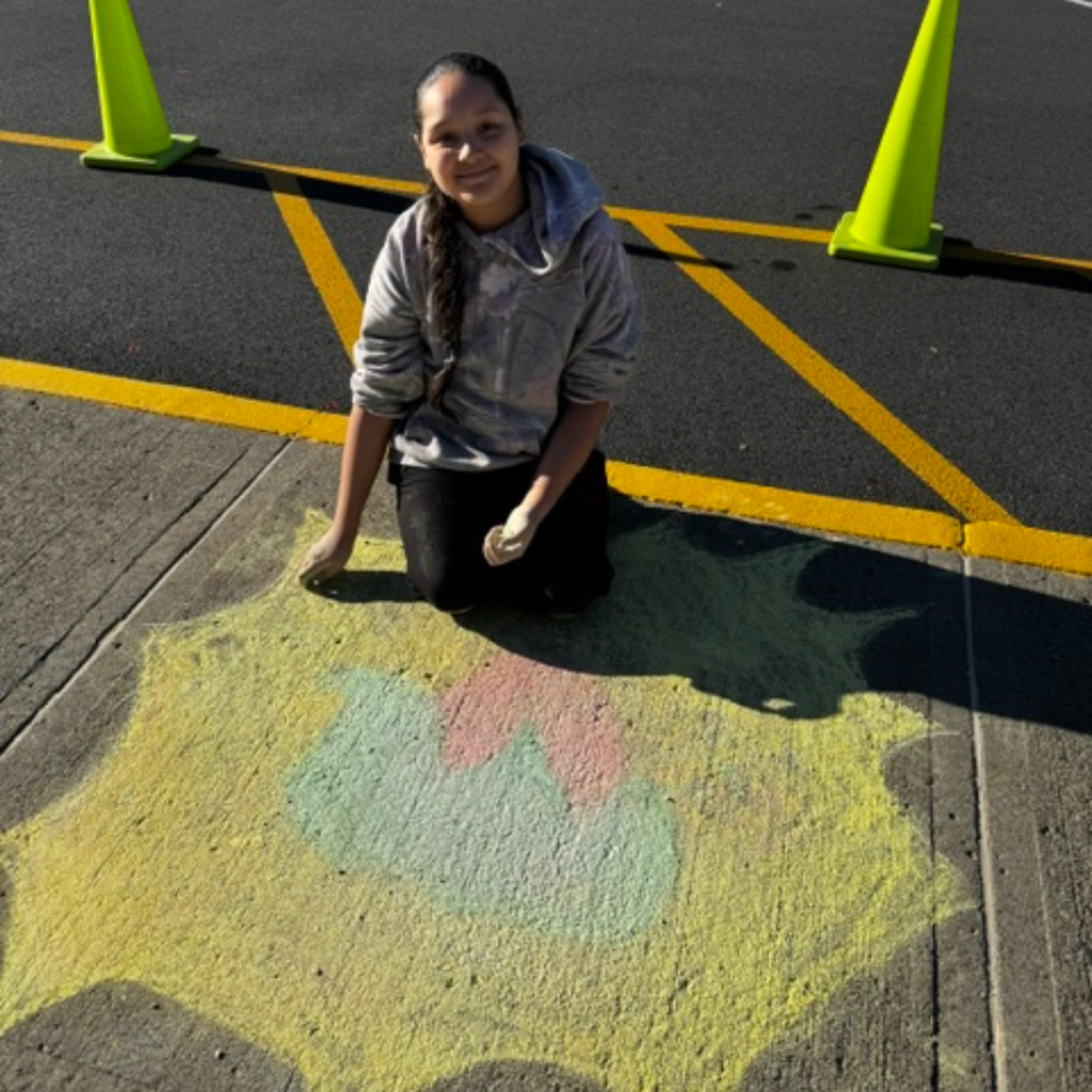 Girl kneeling in parking lot in front of yellow, pink, and green chalk design. Two traffic cones behind.
