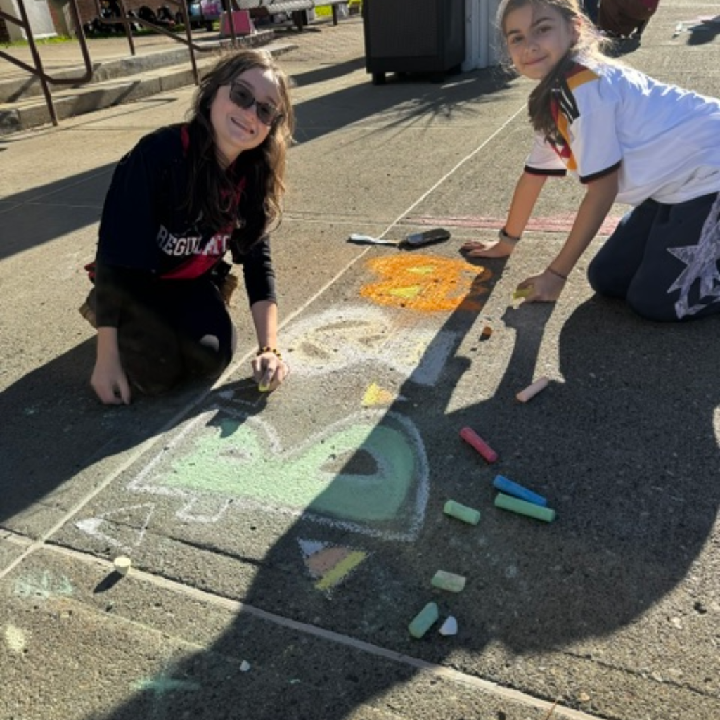 Two people drawing letters and symbols on pavement with chalk, one wearing glasses, other wearing a white shirt.