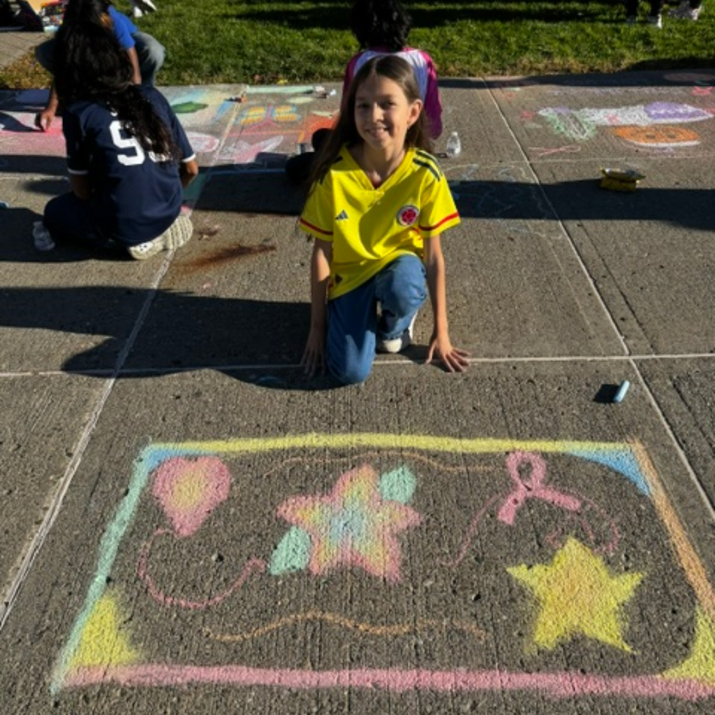 Girl wearing yellow shirt kneeling on sidewalk, drawing with chalk on ground. Other students sitting on ground.
