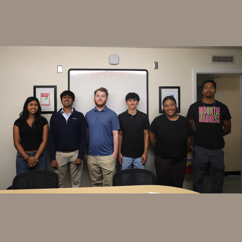 six students standing in a room with a brown table