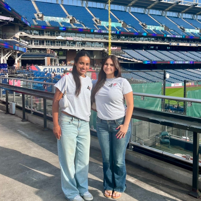 two girls in white t shirts and blue jeans at a baseball park