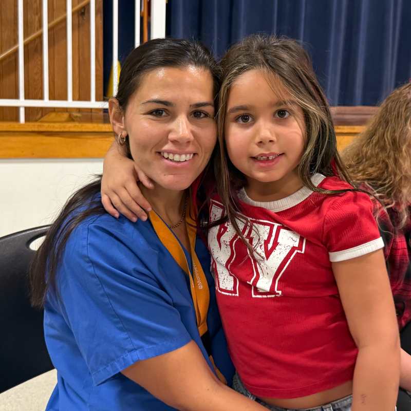 woman with blue shirt and orange tag sits with girl with red shirt