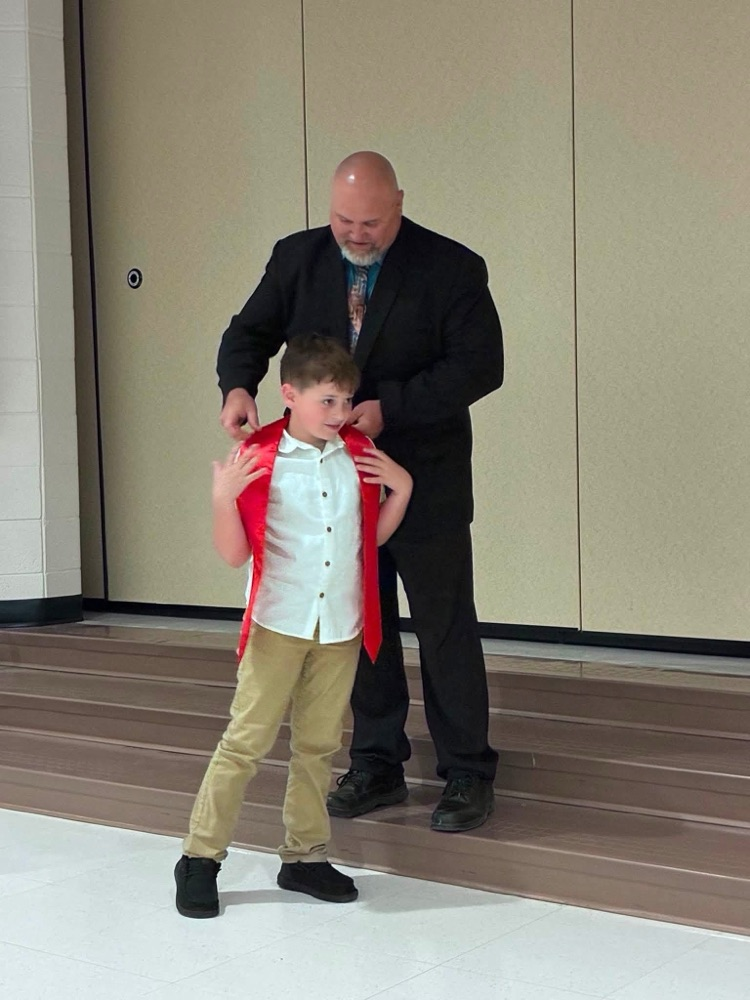 student in tan pants white shirt Jason Conaway in a suit putting on a sash 