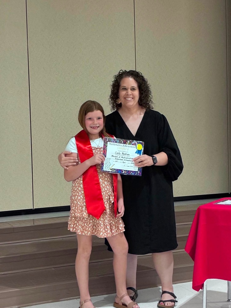 student in a tan and white dress with Miss Willls putting on a red 