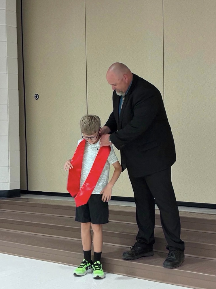 student in gray shorts and gray shirt with Dr. Conaway putting on a red sash 