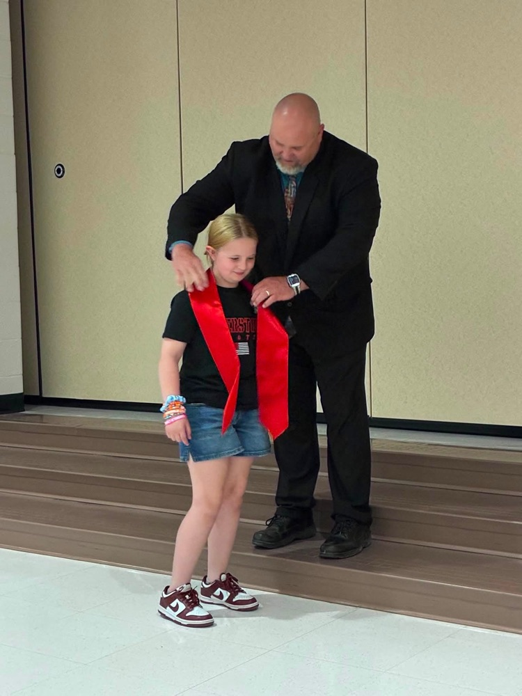 student in blue jean shorts with a black shirt with Dr. Conaway putting on a red sash  