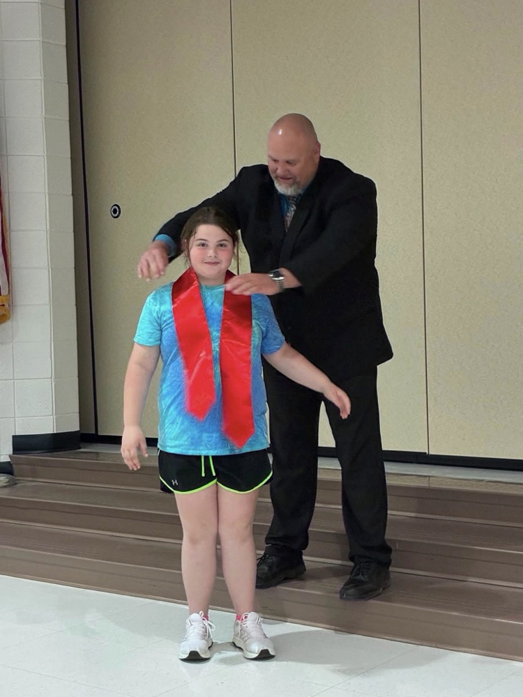 student in a blue shirt and black shorts with Dr. Connaway putting on a red sash