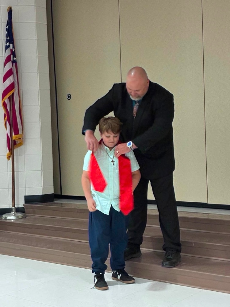 student in green shirt blue pants with Dr. Connaway putting on a red sash