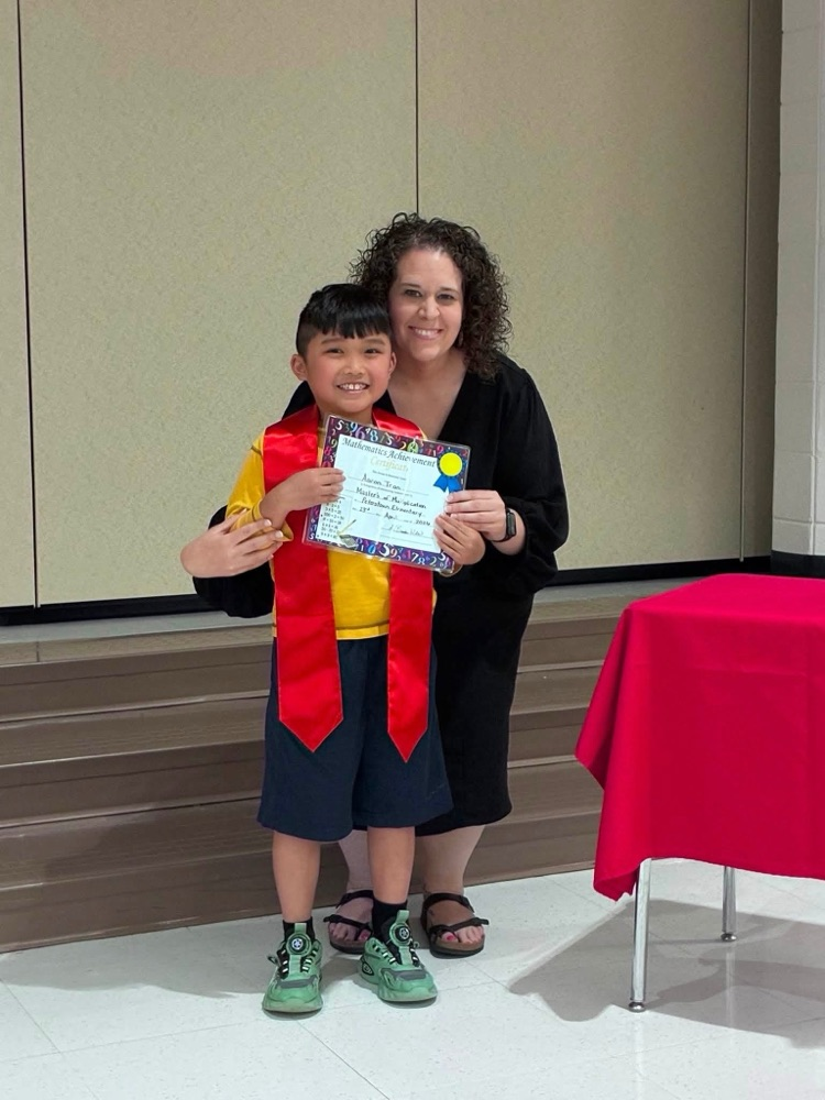 student and yellow shirt blue pants with Miss Wills  putting on a red sash