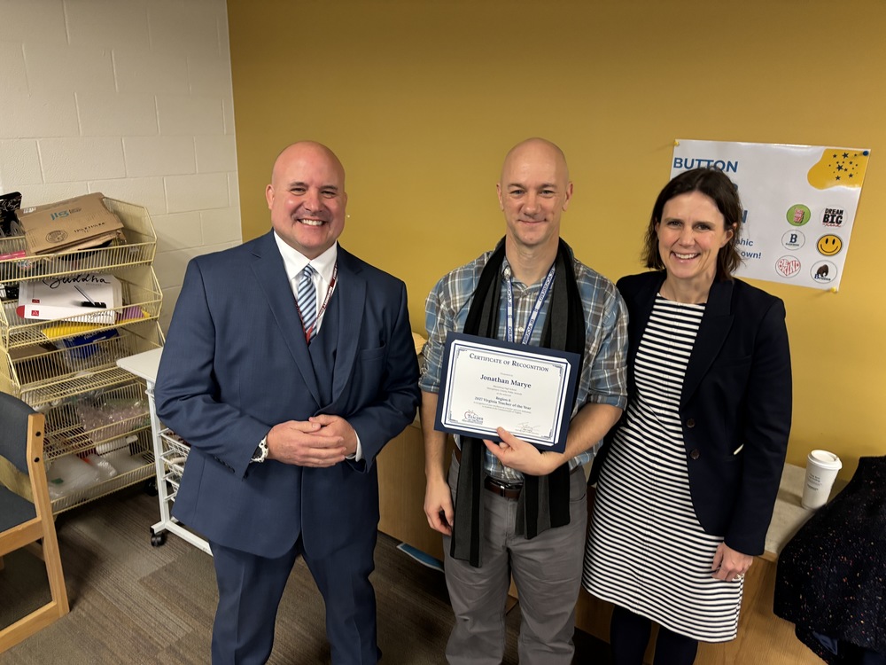 Three people standing together indoors as Jonathan Marye holds a "Region 6 Teacher of the Year" certificate. 