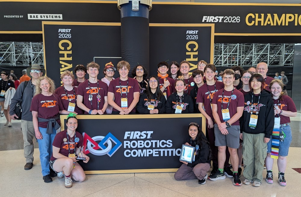 A large group of high school students and adult mentors from FRC Team 401 Copperhead Robotics pose for a photo at the FIRST 2026 Championship. They are gathered around and kneeling behind large black signs that feature the FIRST logo and competition branding. Most are wearing maroon or black Team 401 shirts and hoodies with competition lanyards. A student on the left kneels holding a trophy, while a student on the right kneels holding a framed award. In the background, there is large "FIRST 2026 CHAMPION" branding and an open stadium view.