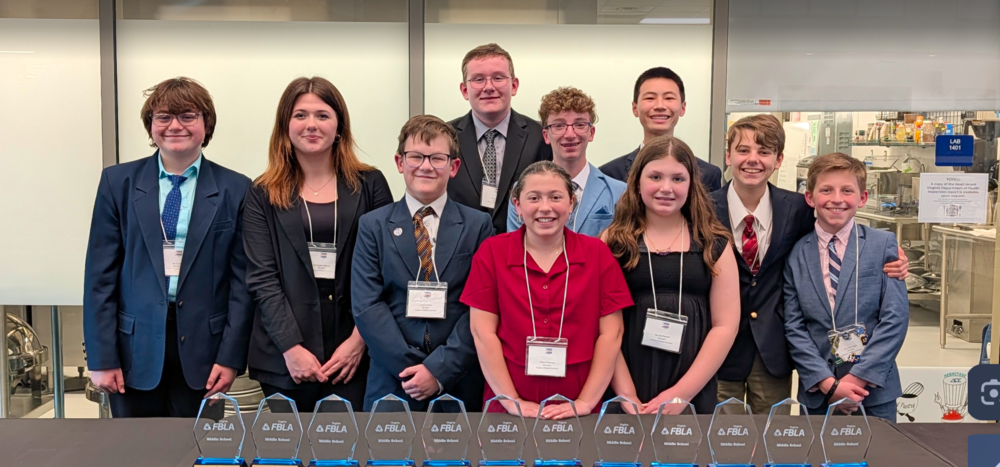 Gemini said A medium outdoor wide-shot photo shows nine students, seven males and two females, as they stand on two different levels and pose as a group. Each male student wears a blazer or suit jacket with slacks while the two females wear nice dresses. The male on the far left wears a dark navy blazer and a light blue dress shirt with a bright teal blue patterned tie and beige slacks. The female next to him wears a black wrap-around style long sleeve dress while the male next to her wears a blue blazer with an off-white button-down shirt and a brown and yellow tie and brown pants. Directly behind him stands a taller student with a dark navy jacket over an off-white shirt and brown tie with beige slacks. The male next to him wears a light blue blazer over a blue and white striped shirt with a navy patterned tie and beige slacks. Beside him is a male student who wears a dark navy blazer with an off-white shirt and a navy tie and beige slacks. The female stands directly in front of him, wearing a dark red, short-sleeve dress and a beaded necklace. To her left is a taller female who wears a black scoop-neck dress and the male on her left wears a navy blazer over a light pink striped shirt with a brown patterned tie. Nine rectangular crystal awards with a pointed top are arranged on a black tablecloth in a horizontal row on the table in front of the group of students. The awards read, from top to bottom, "FBLA" in the middle of a triangle logo with "FBLA" written on each side of the triangle. Written in smaller font below is "Virginia." A row of silver kitchen equipment sits behind the students and they stand on a light-colored tile floor. A yellow and white paper sign is attached to the wall in the right-hand background and reads, "Warning." Small black text follows below. Two vertical blue bars frame the image on the far left and right.
