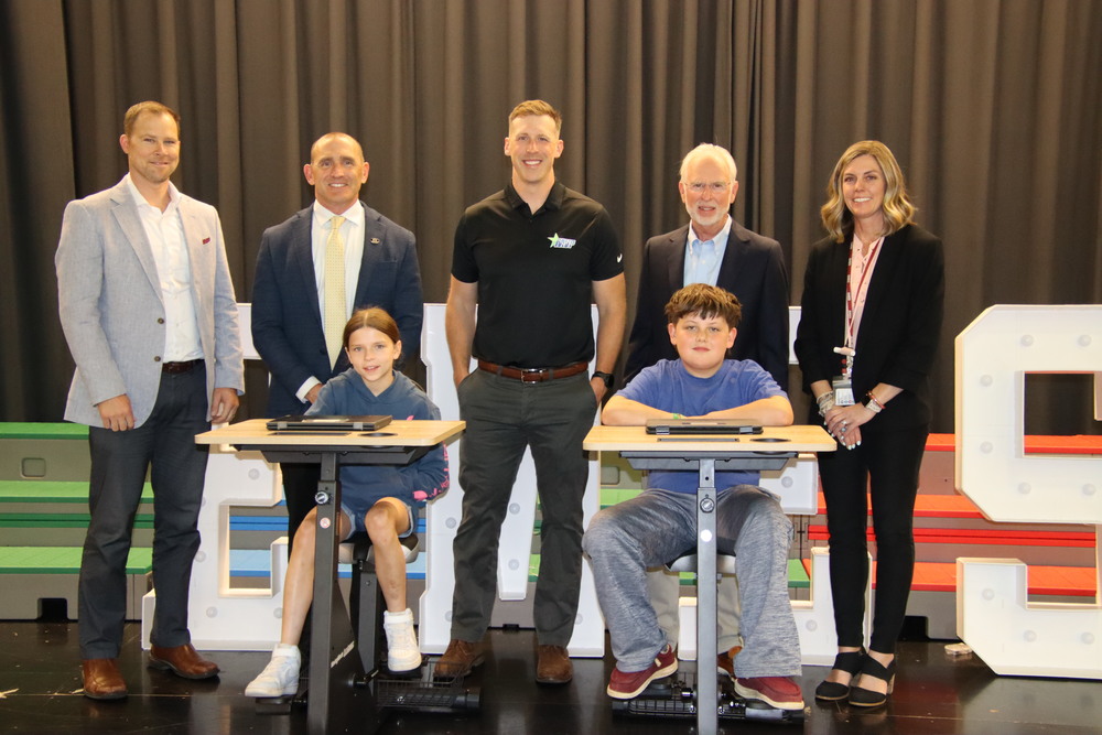 Two EMES students sitting on pedal desks with 5 peoplel standing in the background